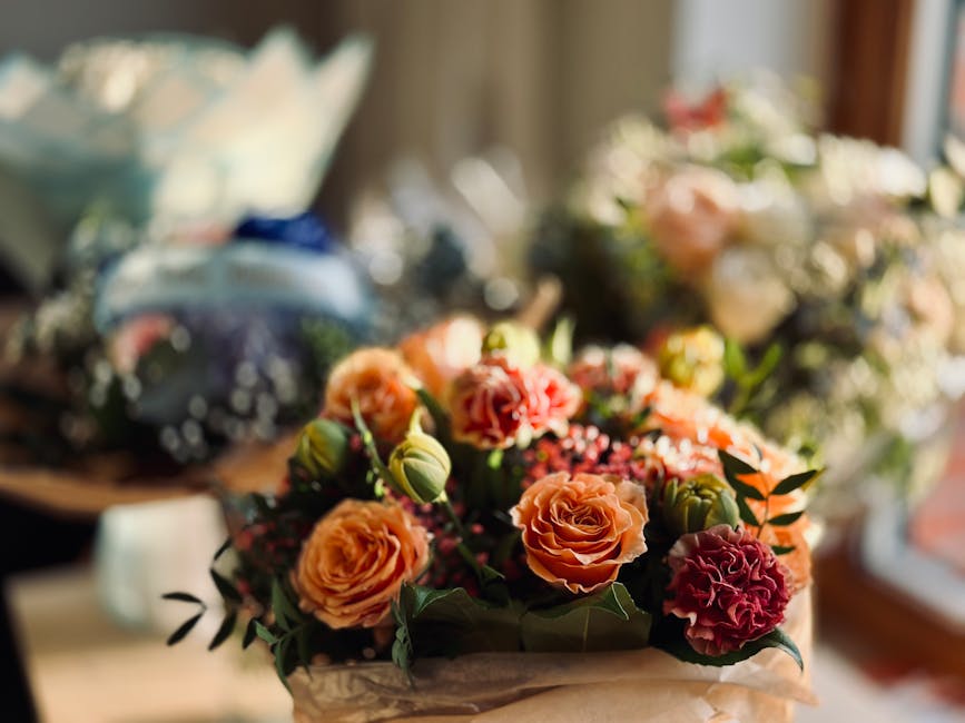 A floral arrangement featuring a bouquet of peach and coral roses, alongside deep red carnations and small pink buds, accented with green foliage and leaves. The flowers are arranged in a natural, lus