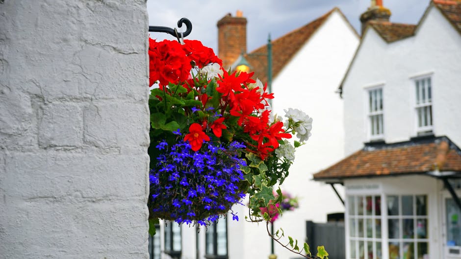 A vibrant hanging floral arrangement attached to a white brick wall, featuring large red geraniums, white petunias, and small blue lobelias, with green foliage. The flowers are arranged in a black wro