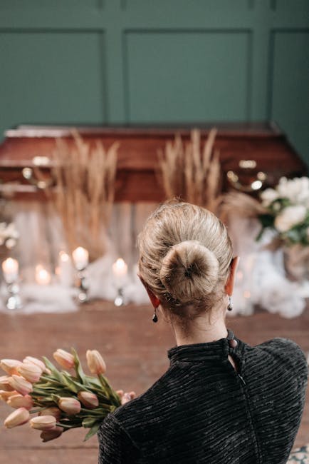 A funeral arrangement featuring a polished wooden coffin surrounded by white and cream-colored roses, anthuriums, and greenery, with tall, beige pampas grass in the center. The arrangement rests on a