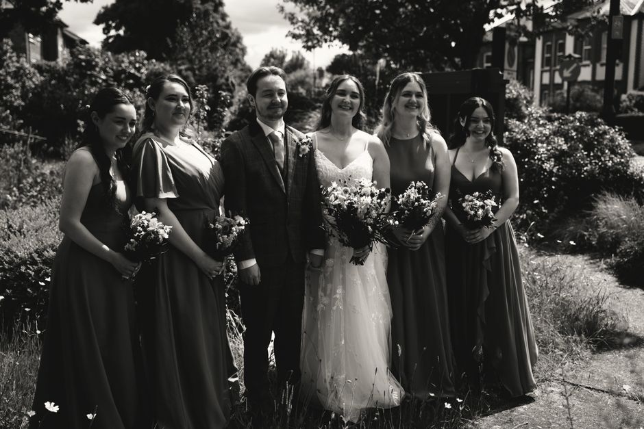A black and white photograph capturing a bride and five bridesmaids outdoors during a wedding ceremony. The bride is at the center, wearing a white lace wedding gown and holding a large bouquet of mix