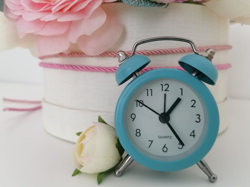 A close-up of a vintage-style light blue alarm clock with two bells on top, showing the time as 12:03, placed in front of a white, cylindrical gift box decorated with pink and white ribbons. To the le